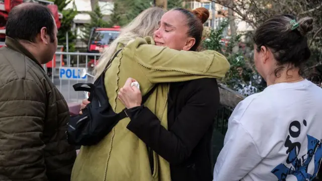 Fotoğrafta, 2 Nisan 2024'te Gayrettepe'deki yangının ardından yakınlarından haberdar olmak üzere mekanın önüne gelen dört kişi görülüyor. İki kadın birbirine sarılarak ağlıyor. Bir adam ve bir kadın ise bekliyor. Arkada polis barikatı görülüyor. 