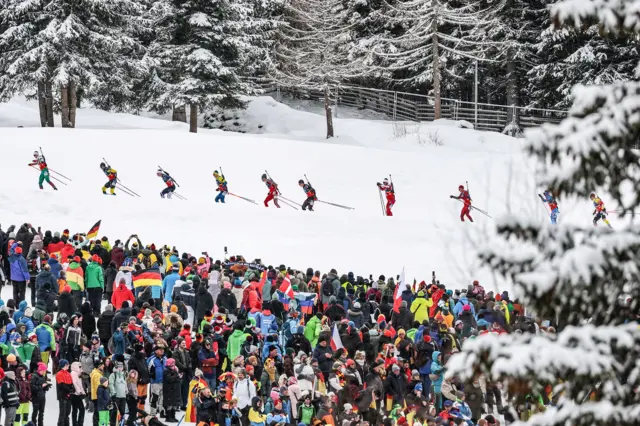 Биатлонисты во время эстафеты, на переднем плане - множество болельщиков в разноцветной одежде 
