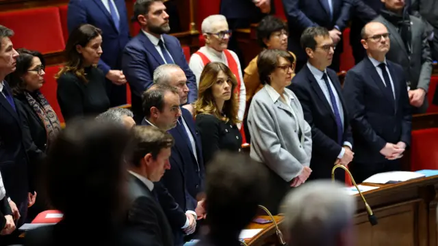 Un grupo de parlamentarios franceses con las manos cruzadas y gesto serio guarda un minuto de silencio