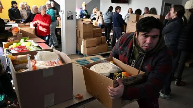 رجل يحمل صندوق طعام في قاعة كنيسة. وهناك صناديق أخرى مكدسة في أرجاء القاعة، والناس يقفون ويعملون في الخلفية.