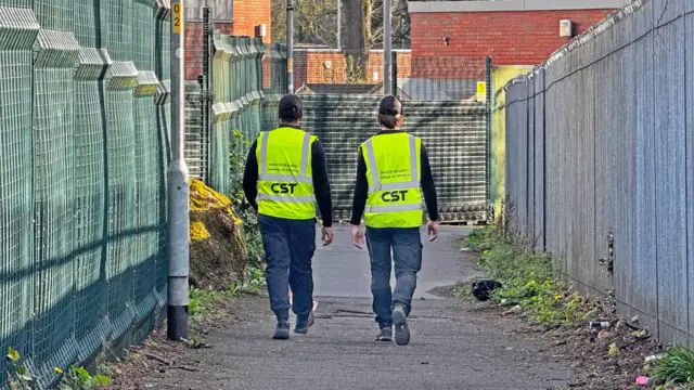 رجلان يرتديان قبعات سوداء وسترات عاكسة للضوء وسراويل داكنة يسيران مبتعدين عن الكاميرا على طول ممر واسع للمشاة، محاط بأسوار عالية من الجانبين.