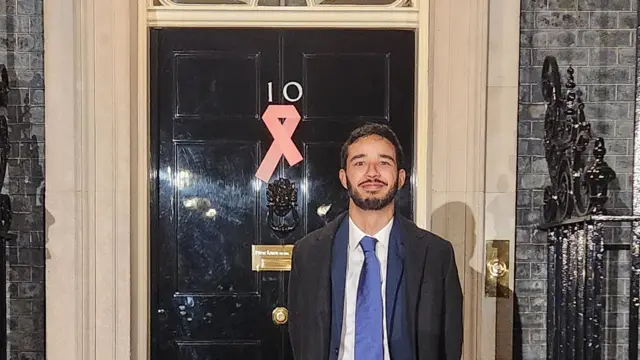 A picture of Hamish standing in front of the door of 10 Downing Street, where there is a red ribbon to symbolise solidarity with people living with HIV 