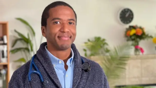 Доктор Мур, темнокожий молодой мужчина с короткой стрижкой, на шее висит стетоскоп.