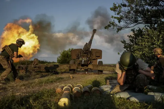 Момент пострілу з великої артилерійської гармати на відкритій місцевості. Із каналу гармати виривається яскравий вогняний спалах і густий дим. Ліворуч військовий у спорядженні тягне трос для здійснення пострілу. Справа двоє військових укриваються та затуляють вуха від звуку. На передньому плані на землі лежить кілька артилерійських снарядів. Довкола — трава, кущі та відкрите небо у вечірньому світлі.