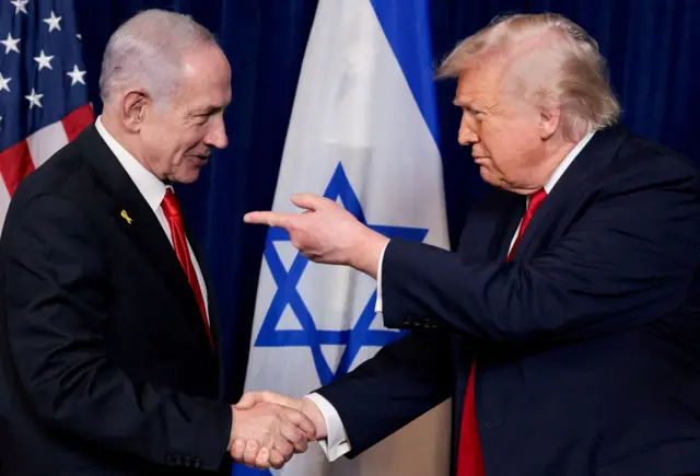 Benjamin Netanyahu ve Donald Trump, ABD ve İsrail bayraklarının önünde dururken el sıkışıyor. Her iki adam da kırmızı kravatlı koyu mavi takım elbise giyiyor, Trump ise Netanyahu'yu işaret ediyor.