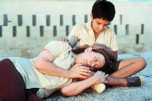 A atriz Fernanda Montenegro aparece deitada no chão, com expressão abatida e olhar distante, enquanto o ator Vinícius de Oliveira está sentado atrás dela, apoiando a mão sobre sua cabeça. Ela veste uma blusa clara, enquanto ele usa camisa bege e shorts escuros. O cenário ao fundo é árido, com postes fincados na paisagem seca.