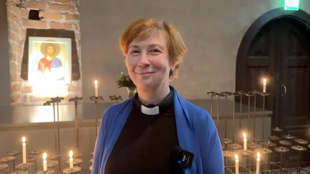 مکنین اولیکی در مقابل شمع‌های فروزان و تصویری از مسیح ایستاده است. او یک یقه مخصوص کشیش‌ها و یک ژاکت کش باف پشمی آبی پوشیده است. او موهای کوتاه وسرخ‌رنگ دارد