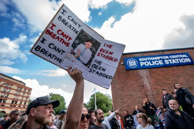 Grupa ludzi protestujących przed komisariatem policji w Southampton Central. Jedna osoba trzyma transparent ze zdjęciem Henry'ego Nowaka, a pod nim napis „Zostałem dźgnięty nożem” i „Nie mogę oddychać”.
