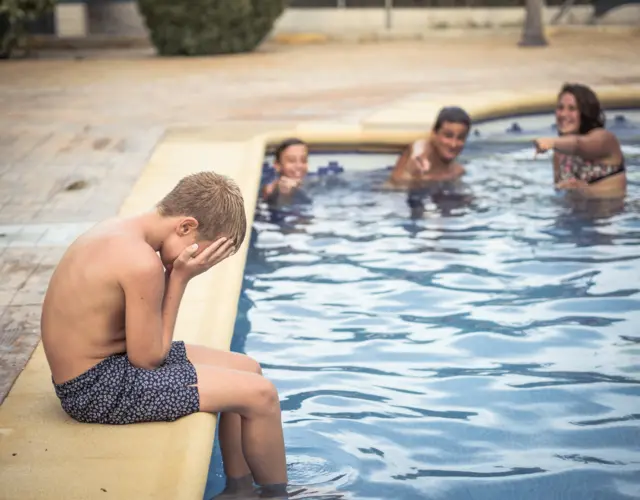 Criança branca chorando na piscina, no fundo, três pessoas brancas riem