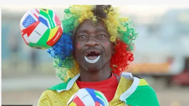 Ọkunrin to de irun pẹlu ọda awọ ọlọkan-o-jọkan lori, bọọlu kan wa nibi ori rẹ, o lanu pẹliu eyin rẹ to pa eji nla.