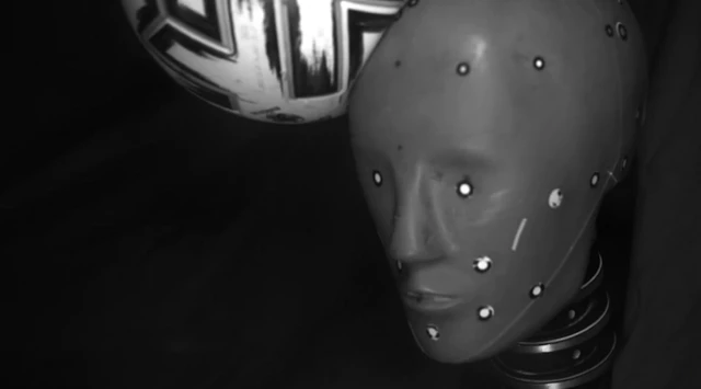 Сива глава лутке са белим сензорима на различитим деловима лица, коју је ударила бело-црна фудбалска лопта са десне стране