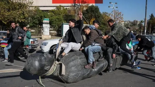 چند جوان روی مجسمه سرنگون شده حافظ اسد، رئیس جمهور پیشین نشسته‌اند و شادی می‌کنند
