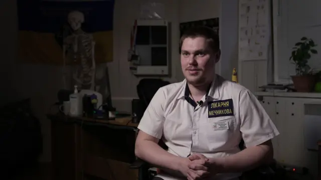 Максим Макаренко, травматолог, сидить на тлі скелета у медичній сорочці білого кольору з написом "Лікарня Мечникова"