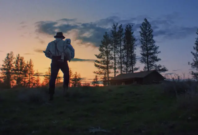 A imagem mostra a silhueta de um homem caminhando por um campo ao entardecer, carregando nos braços uma criança envolta em um pano claro. Ele usa chapéu, camisa de mangas compridas com suspensórios e calças escuras. Ao fundo, o céu está tingido por tons de laranja e azul do pôr do sol, com nuvens espalhadas e a silhueta de árvores altas. À direita, há uma casa de madeira simples, parcialmente envolta pela penumbra, reforçando a atmosfera rural e contemplativa da cena.