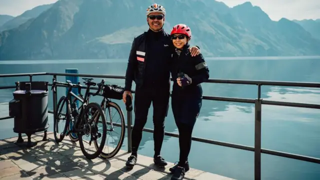 Suami-istri Devi dan Budiman berpose di depan danau saat liburan berdua