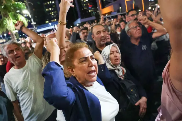 Kalabalık bir protesto. Orta yaşlı kadınlar ve erkekler ellerini yumruk yapmış bağırıyor.