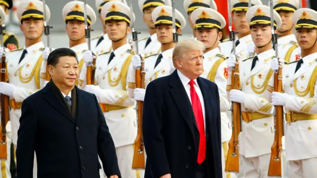 Şi Jinping ve Donald Trump beyaz giyinmiş ve silah tutan Çin birliklerinin yanından geçiyor. Şi ve Trump koyu renk takım elbise giyiyor.
