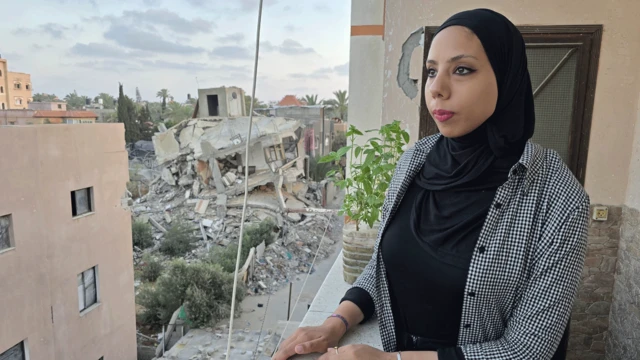 Hanya Aljamal olha para o horizonte, da sacada de sua casa. Seu cabelo está coberto por um lenço preto. Ao fundo, podem-se ver edifícios destruídos.