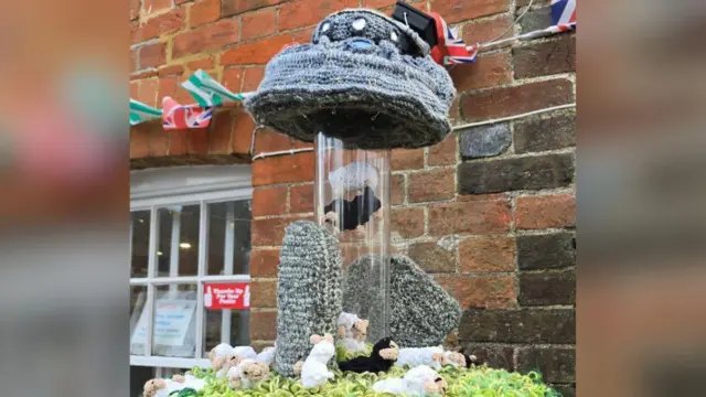 Un adorno para buzón, hecho de "hierba" de lana verde con varias figuras de ovejas blancas y negras esparcidas. Hay un tubo de plástico entre dos piedras verticales tejidas a ganchillo, y en él hay tres figuras de ovejas que parecen ser recogidas por un platillo volador gris también tejido a ganchillo. El buzón está frente a la oficina de correos del pueblo, un edificio de ladrillo rojo con banderines.