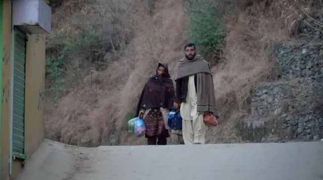 زن و مردی در خیابانی در روستای پیرا در کنار هم راه میروند و کودکی نیز در میانشان دیده میشود. زن و مرد لباسهای سنتی به تن دارند و هر دو خرید روزانهشان در دستهایشان است. مرد در حال نگاه کردن به لنز دوربین است