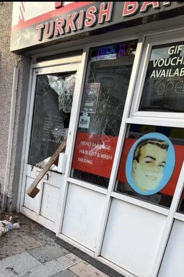 Saldırıya uğrayan Türk berberinin camları kırılmış. Tabelada "Turkish Barber" yazıyor. Dükkanın camında traş olan bir adam çizimi var.