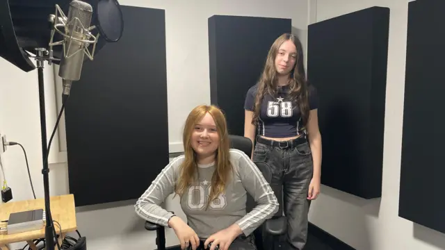 Bethany Quinn está sentada en el estudio de grabación, con una camiseta gris de manga larga con rayas blancas. La camiseta dice 08 y tiene una estrella sobre los números. Tiene el pelo largo y rojizo, y el micrófono plateado está a la izquierda, sobre un soporte negro. Elsie Williamson está a su derecha, ligeramente detrás. Lleva una camiseta negra corta con el número 58, también con una estrella sobre él. Lleva vaqueros grises y un cinturón negro.