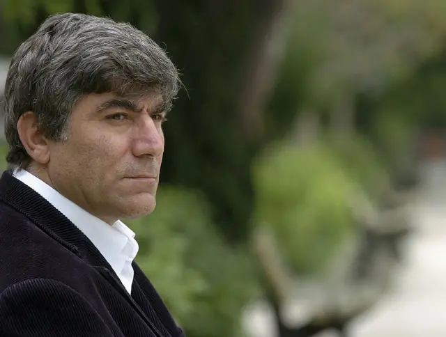 Esmer bir adam uzaklara bakıyor. Fonda bir bahçe ve banklar var.