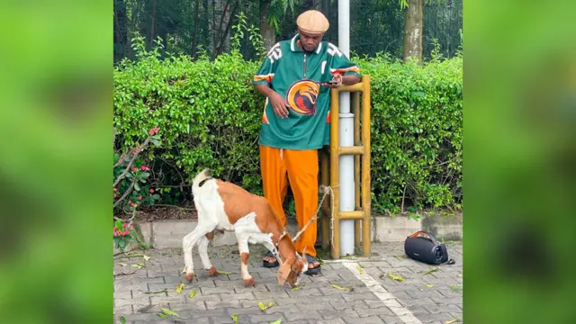 प्रॉमिस नारंगी पतलून और हरे रंग की टॉप पहने एक लैंपपोस्ट के पास खड़ा है. उसके बगल में एक बकरा लैंपपोस्ट से बंधा हुआ है.
