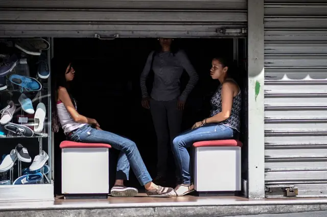 Empleadas de una zapatería aparecen en una fotografía sentadas a la entrada de un local a oscuras durante un corte de electricidad en Caracas.