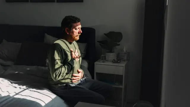Egy férfi ül egy ágy szélén, egyik keze a mellkasán, a másik a hasán, miközben a napfény az arcára süt az ablakon keresztül.