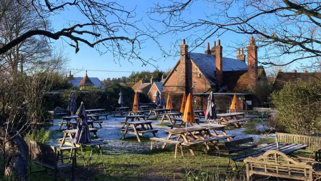 Una escena en el jardín de un pub con una ligera capa de nieve entre las mesas de picnic y las sombrillas. El jardín está desierto con un cielo azul brillante.