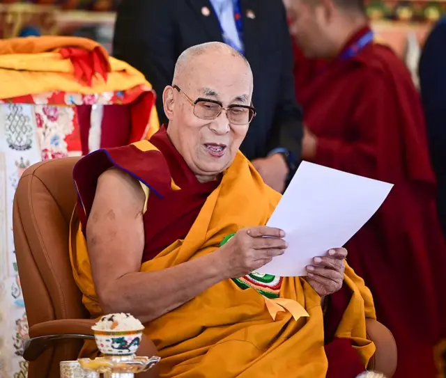 Dalai Lama geleneksel kıyafetiyle oturuyor ve elinde beyaz bir kağıt tutuyor.