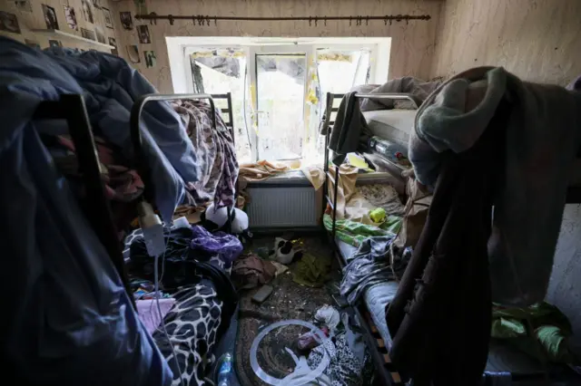 Заваленная вещами комната с двумя двуспальными кроватями по бокам, по центру полуразрушенное окно
