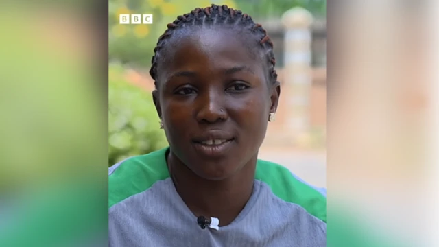 A youn woman wit braided hair, nose ring and earring. She open her mouth wit her teeth showing. She wear ash cloth wey get green line for di shoulder