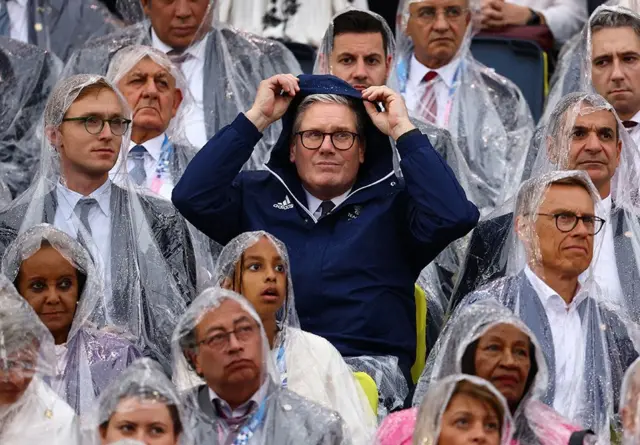 Le Premier ministre britannique Keir Starmer brandit la capuche de son imperméable, entouré d'autres personnes en poncho assises dans les tribunes.
