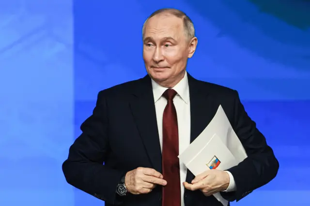Владимир Путин в темном костюме и бордовом галстуке с белыми папками в руке поправляет пиджак