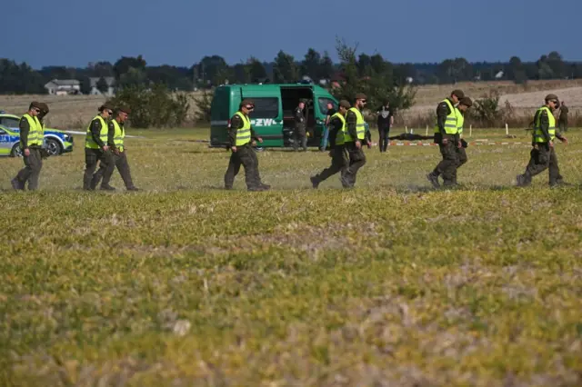 Люди в жёлтых отражающих жилетах оцепляют поле. На заднем плане видны машины польской полиции и спецслужб, а также красно-белая лента ограждения