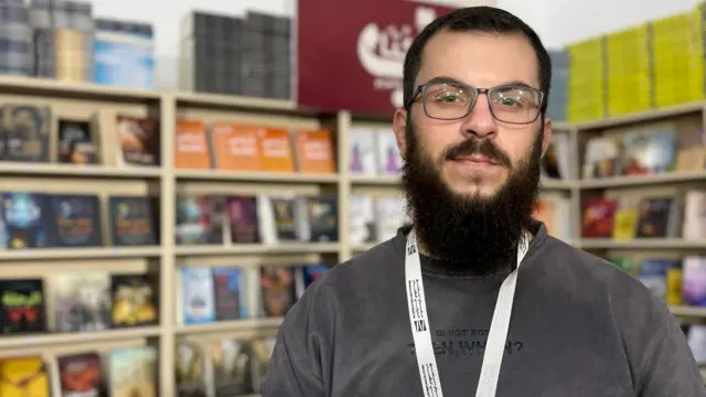 رجل ذو لحية كثيفة وشارب ويرتدي نظارات طبية يقف أمام أرفف معروض عليها كتب مختلفة