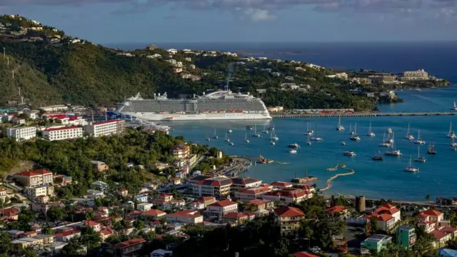Panorama atual do porto de Saint Thomas, com pequenas embarcações e um grande navio de cruzeiro em um mar azul e um céu igualmente azul.