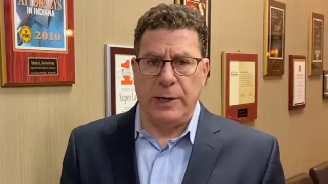 Mark S Zuckerberg arimo kuvuga areba muri 'camera'. Yambaye ikositimu y'umukara, ishati y'ubururu bwerurutse n'indorerwamo. Iruhande rwe hari urukuta ruriho amakadere yanditseho amagambo.