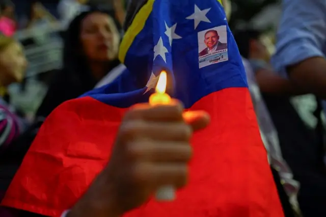 Uma pessoa com uma vela acesa, com o rosto coberto pela bandeira da Venezuela e segurando uma fotocassino que deposita 5 reaisEdmundo González