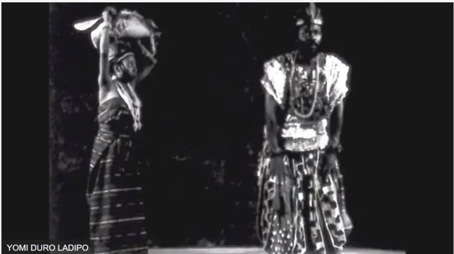 Duro Ladipo ati obinrin kan lori ere ori itage, obinrin naa ru ẹru sori, Duro Ladipo fi ọwọ rẹ mejeeji sori orunkun