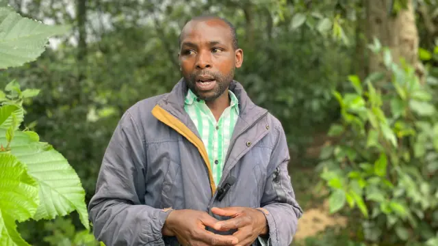 Umugabo uhagaze ahantu hari ibimera byinshi bitoshye, yambaye ikoti ry'ibara rijya gusa n'ivu, ririho indangururamajwi nto, n'ishati irimo ibara ry'icyatsi kibisi n'umweru. 