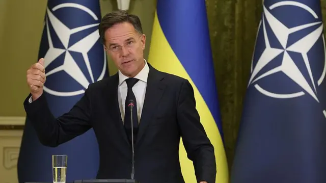 Марк Рютте что-то говорит, жестикулируя, на фоне флагов Украины и НАТО