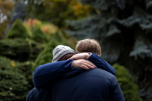 Женщина в сером капюшоне на голове крепко обнимает подростка на фоне зеленого парка, лица обоих скрыты