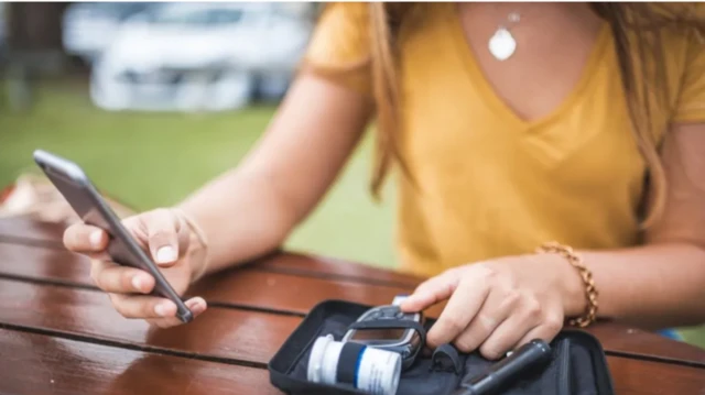 ਮੋਬਾਈਲ ਦੇਖ ਰਹੀ ਕੁੜੀ ਦੇ ਹੱਥ ਇੱਕ ਹੱਥ ਵਿੱਚ ਡਾਇਬਿਟੀਜ਼ ਮਾਨੀਟਰ ਵੀ ਫੜਿਆ ਹੈ