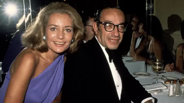 Barbara Walters y Alan Greenspan son fotografiados en una cena de etiqueta en 1977. Miran a la cámara y sonríen. Walters lleva un vestido azul y Greenspan viste un esmoquin.