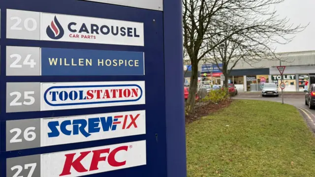 Primer plano de un cartel con una lista de establecimientos fuera de un polígono comercial. Las tiendas se ven al fondo.