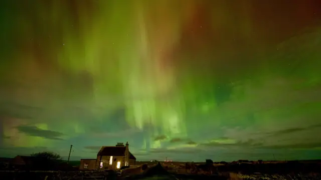 Aurora boreal con vibrantes tonos verdes y rojizos iluminando el cielo nocturno sobre una casa rural y una carretera.