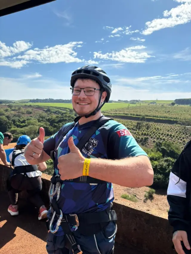 Foto do irmão de Rodrigo, um rapaz jovem, de capacete e equipamentos de salto, fazendo jóia para a câmera. 